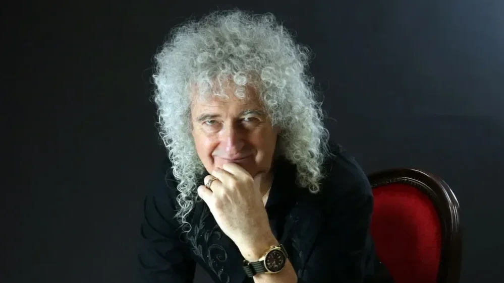 A man with curly gray hair sits and rests his chin on his hand, wearing a black shirt and a gold watch, in front of a dark background.