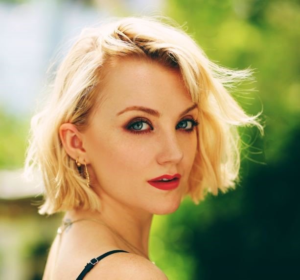 A young woman with blonde hair and blue eyes outdoors, looking at the camera.