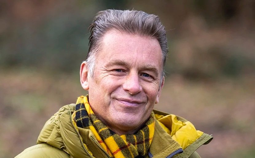 A middle-aged man with gray hair smiling outdoors, wearing a yellow jacket and a black and yellow checkered scarf.