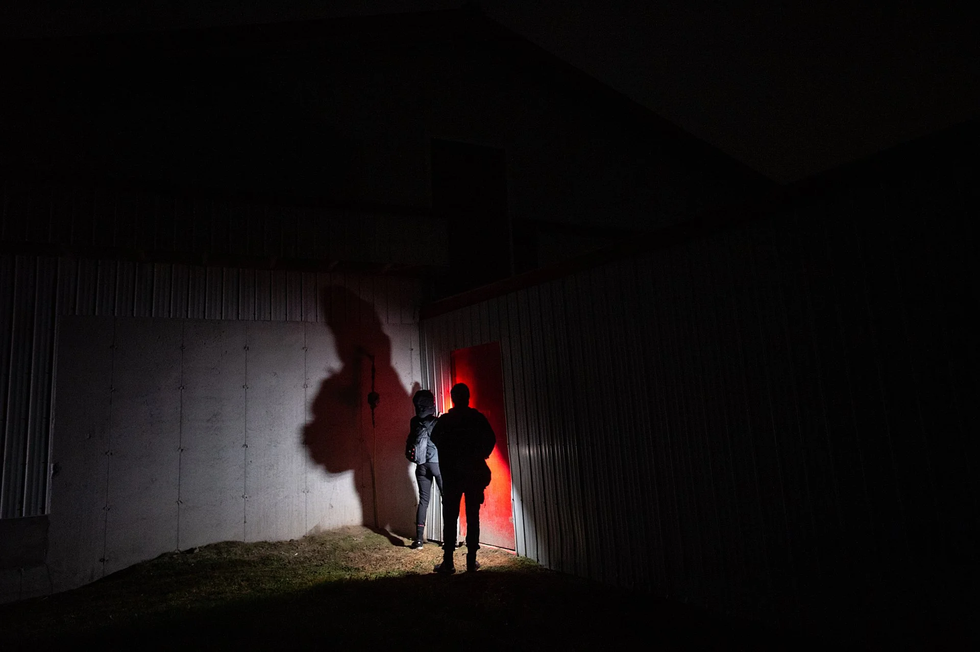 Two undercover investigations are standing outside a facility at night, illuminated by a red light. The surrounding area is dark, with a metal building wall.