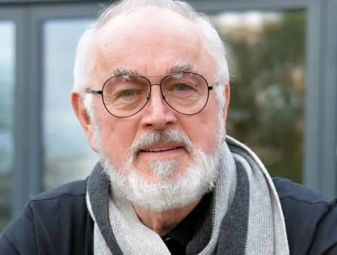 Close-up of an older man with white hair and beard wearing glasses, a gray scarf, and a black jacket, standing outdoors.
