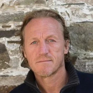 A middle-aged man with light brown hair and stubble, wearing a black zip-up jacket, standing outdoors against a stone wall.