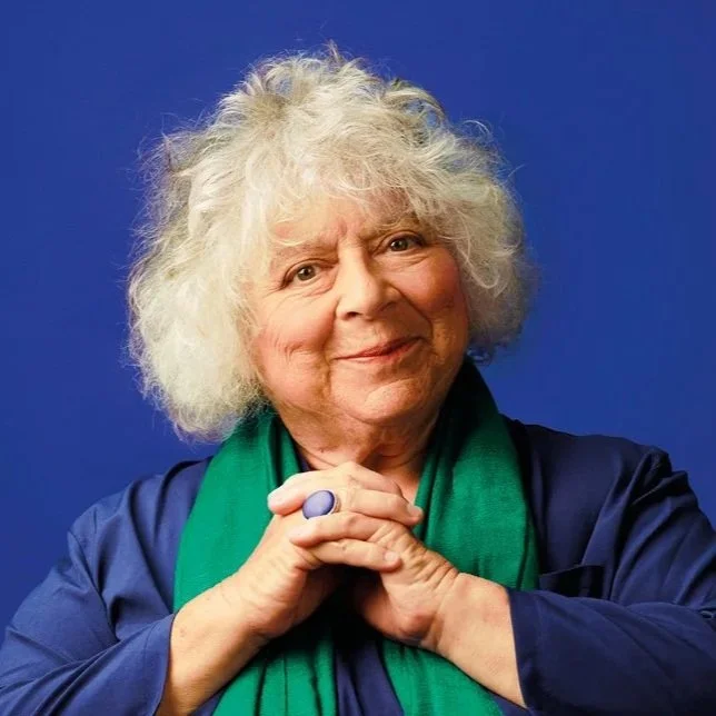 Portrait of an elderly woman with curly white hair, smiling softly, wearing a dark blue jacket and a green scarf, posing with hands clasped against a blue background.
