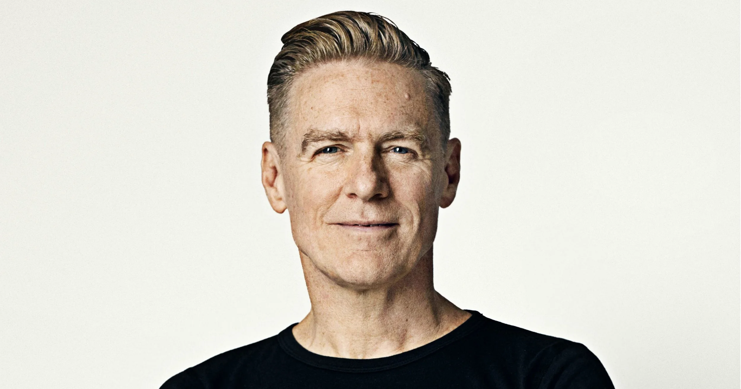 Close-up portrait of a man with light skin, short blonde hair, blue eyes, and wearing a black shirt, smiling gently against a plain white background.