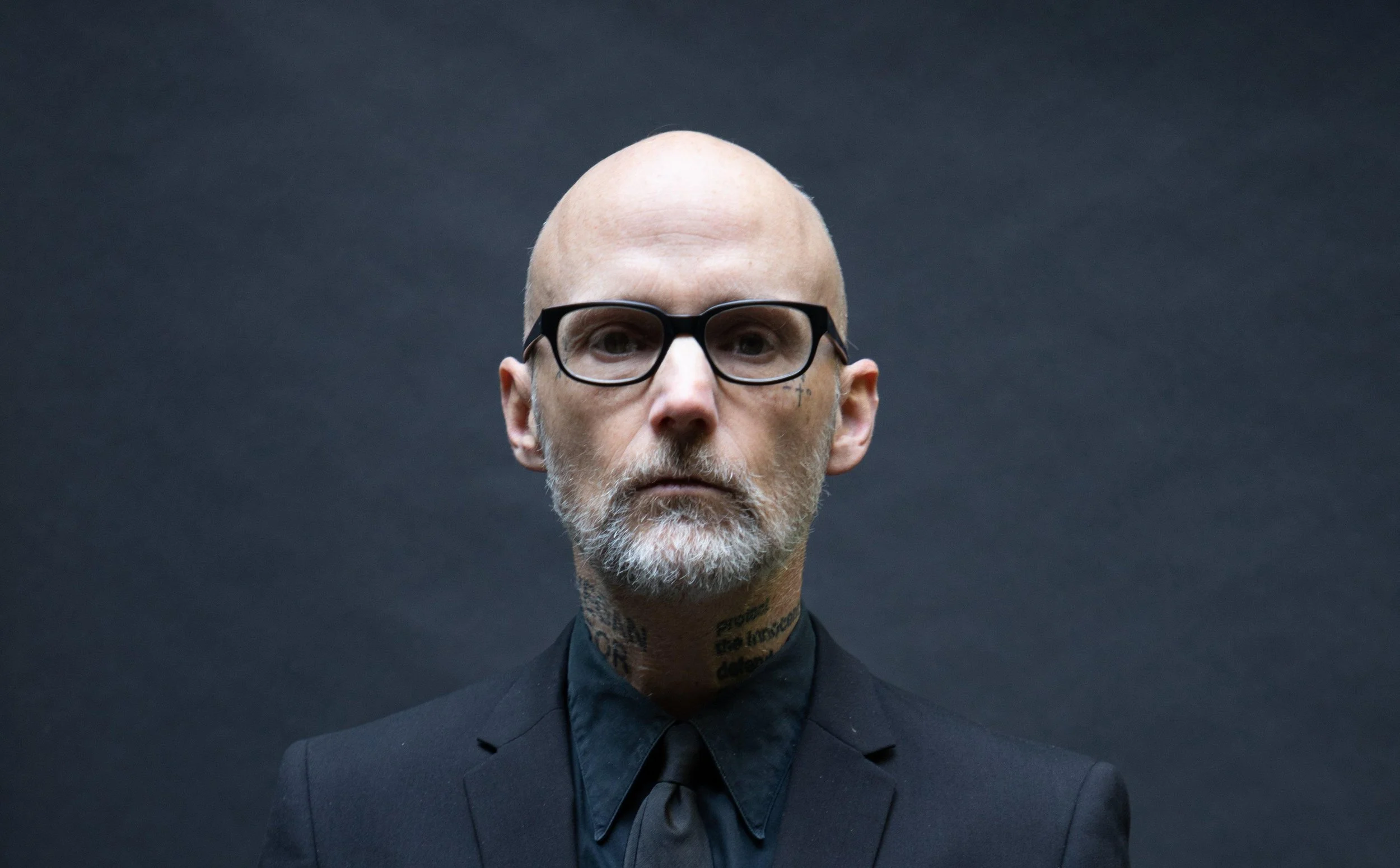 A man with a bald head, glasses, and tattoos on his neck and face, dressed in a dark suit and black shirt, against a dark background.