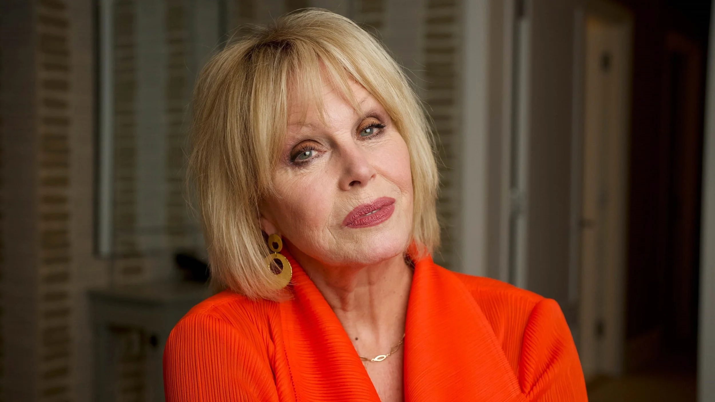 A middle-aged woman with blonde hair and blue eyes, wearing an orange suit and gold earrings, looking at the camera indoors.