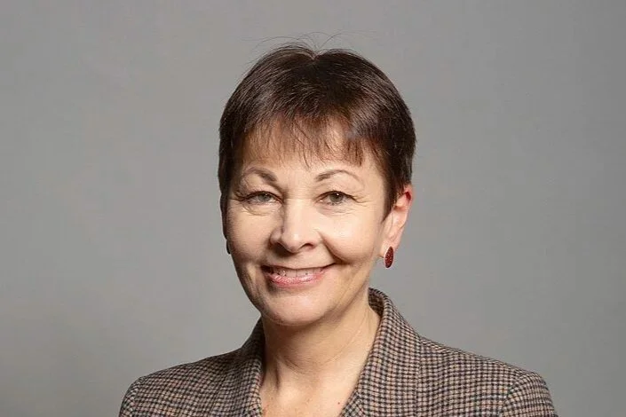 A middle-aged woman with short brown hair, wearing a checked blazer and red earrings, smiling at the camera against a gray background.