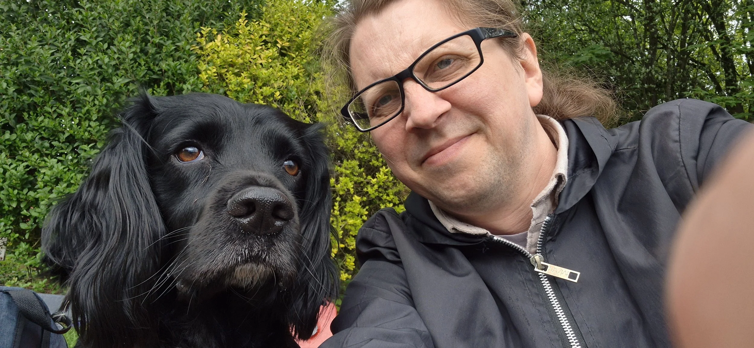A man with glasses taking a close-up selfie with a black dog outdoors with green foliage in the background.