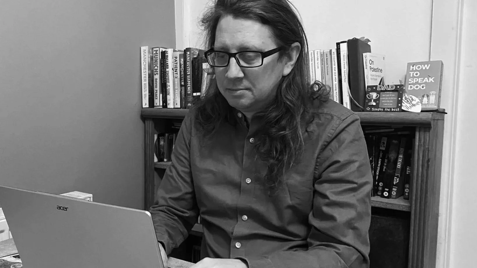 A man with long hair and glasses working on a laptop in a room with a bookshelf behind him filled with books and a couple of children's books on top.