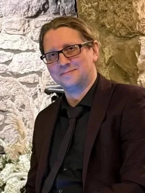 A man with glasses and brown hair, dressed in a black suit and tie, smiling at the camera, in front of a stone wall background.