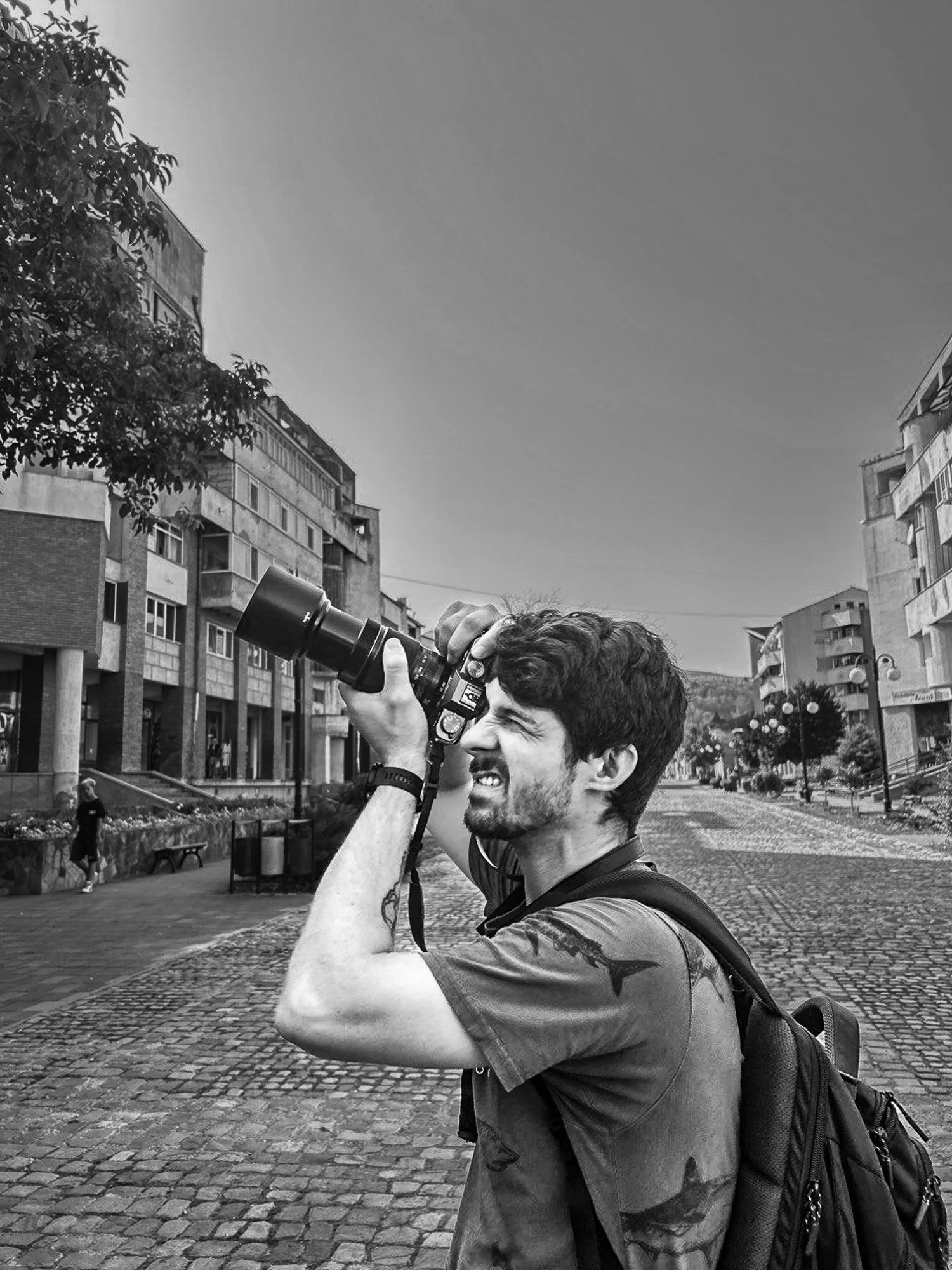 Luka with a beard and tattoos on his arm is taking a photo with a camera in an urban setting with cobblestone streets and multi-story buildings.