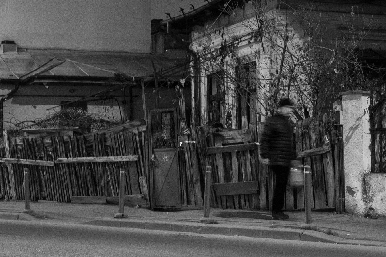 A person walking down on a sidewalk in front of a wooden fence and a building with leafless vines, at night.