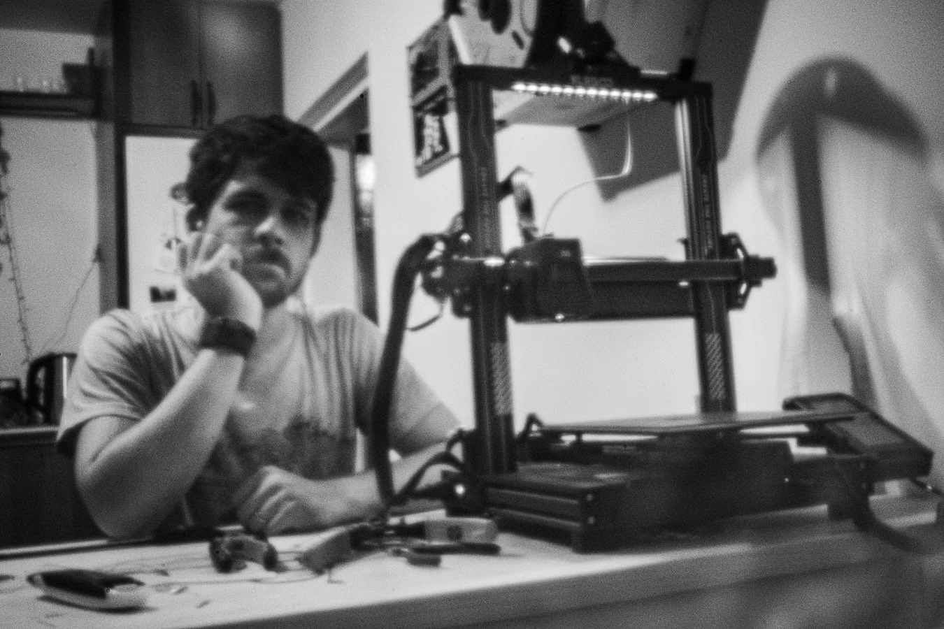 A Luka with dark hair sits at a table with his chin resting on his hand, next to a 3D printer. The room features cabinets in the background and various tools and objects on the table. The image has a dull worn look as it's taken by a pinhole lens.