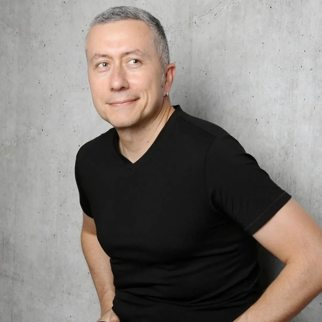 A man with short gray hair, wearing a black t-shirt, standing against a concrete wall. He has a slight smile and is looking to his left.