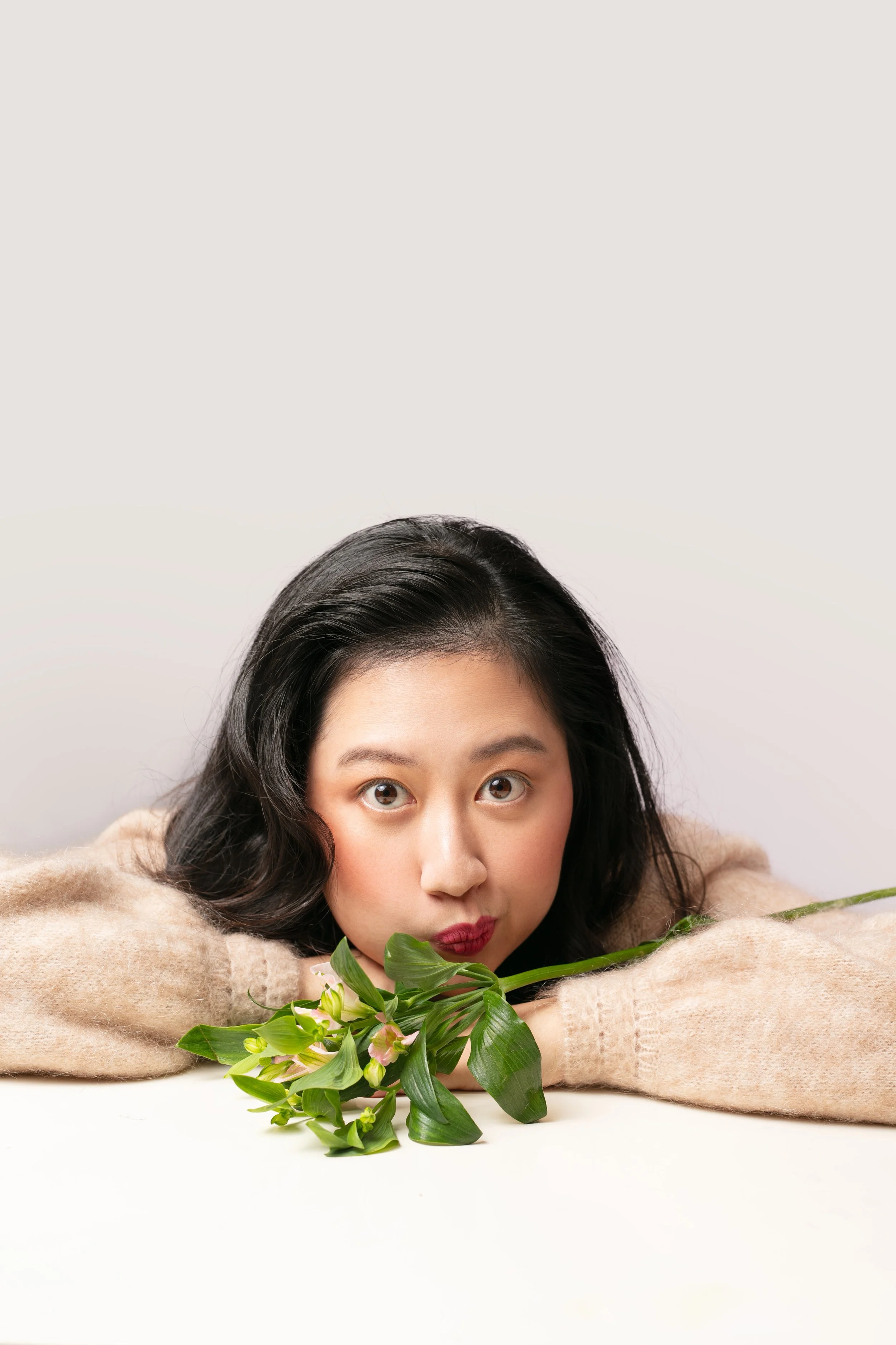 A woman with black hair, wearing a beige sweater, resting her head on her arm and holding a bouquet of green leaves and small pink flowers, against a neutral background.