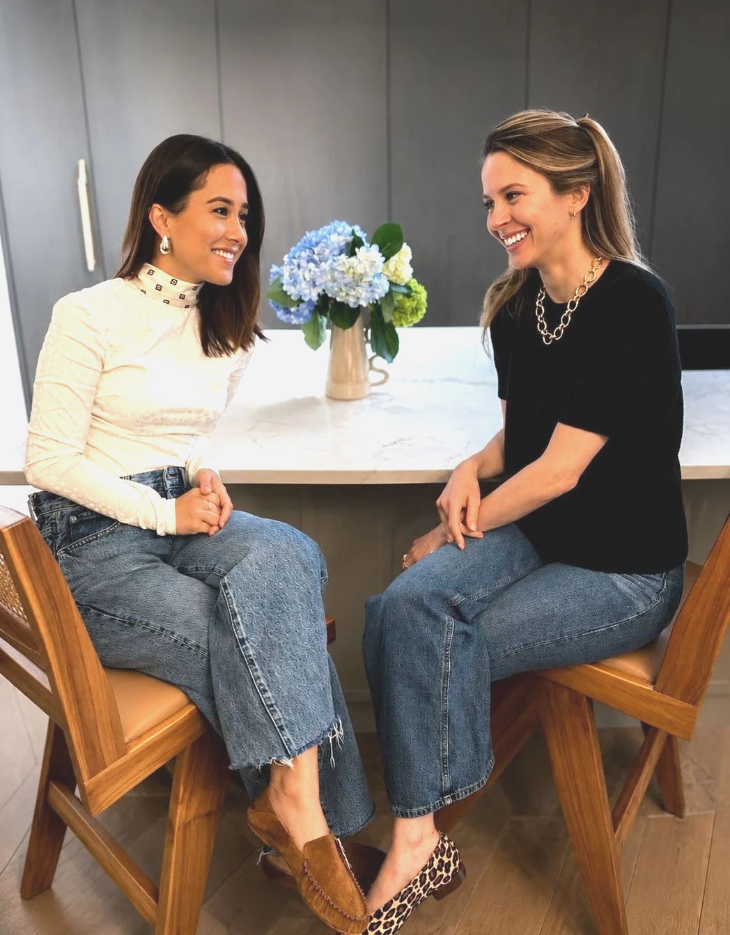 Two women sitting on wooden chairs, facing each other and smiling, with a kitchen counter and a vase of blue and white flowers in the background.