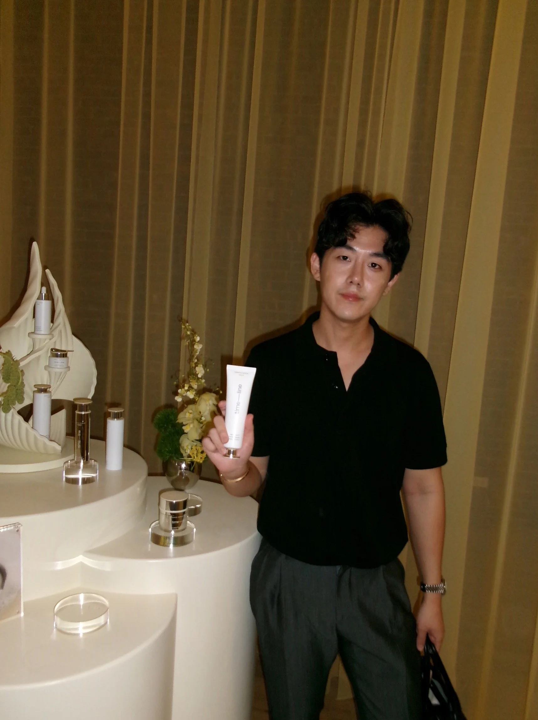 Brute Choi wearing a black shirt and gray pants, standing next to a display of skincare products on a white curved table, holding a tube of skincare product at a promotional event for Timeline