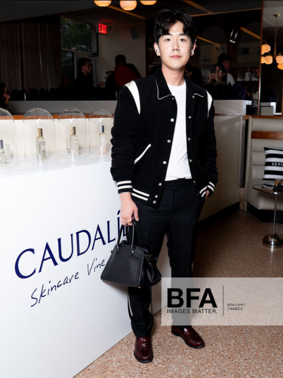Brute Choi wearing a black and white varsity jacket, white shirt, black pants, and red shoes, holding a black handbag, standing indoors at a promotional event for Caudalie skincare.