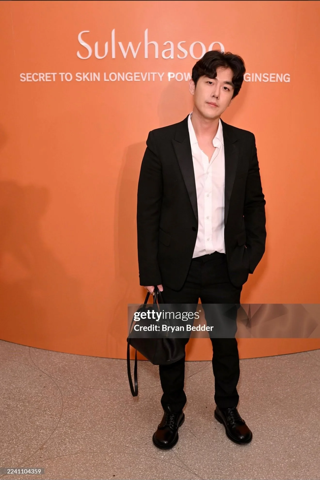 Brute Choi in a black suit and white shirt standing in front of an orange background at a promotional event for Sulwhasoo and their Ginseng line.