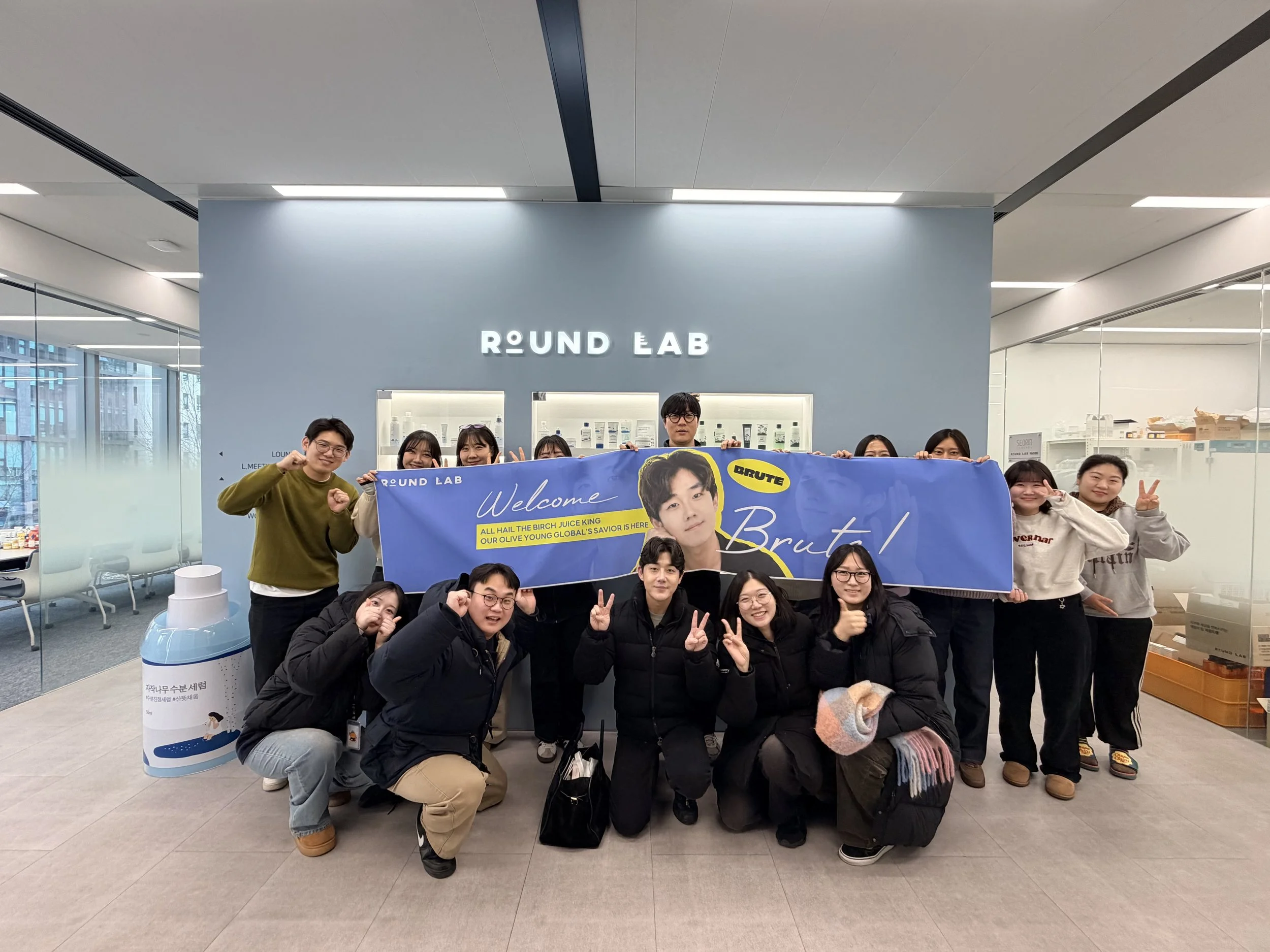 Brute Choi visiting Round Lab headquarters being greeted by a group of people holding a large blue banner in a modern office lobby. Part of Brute's campaign and promotion of Round Lab, specifically the Birch Juice Moisturizing Cream