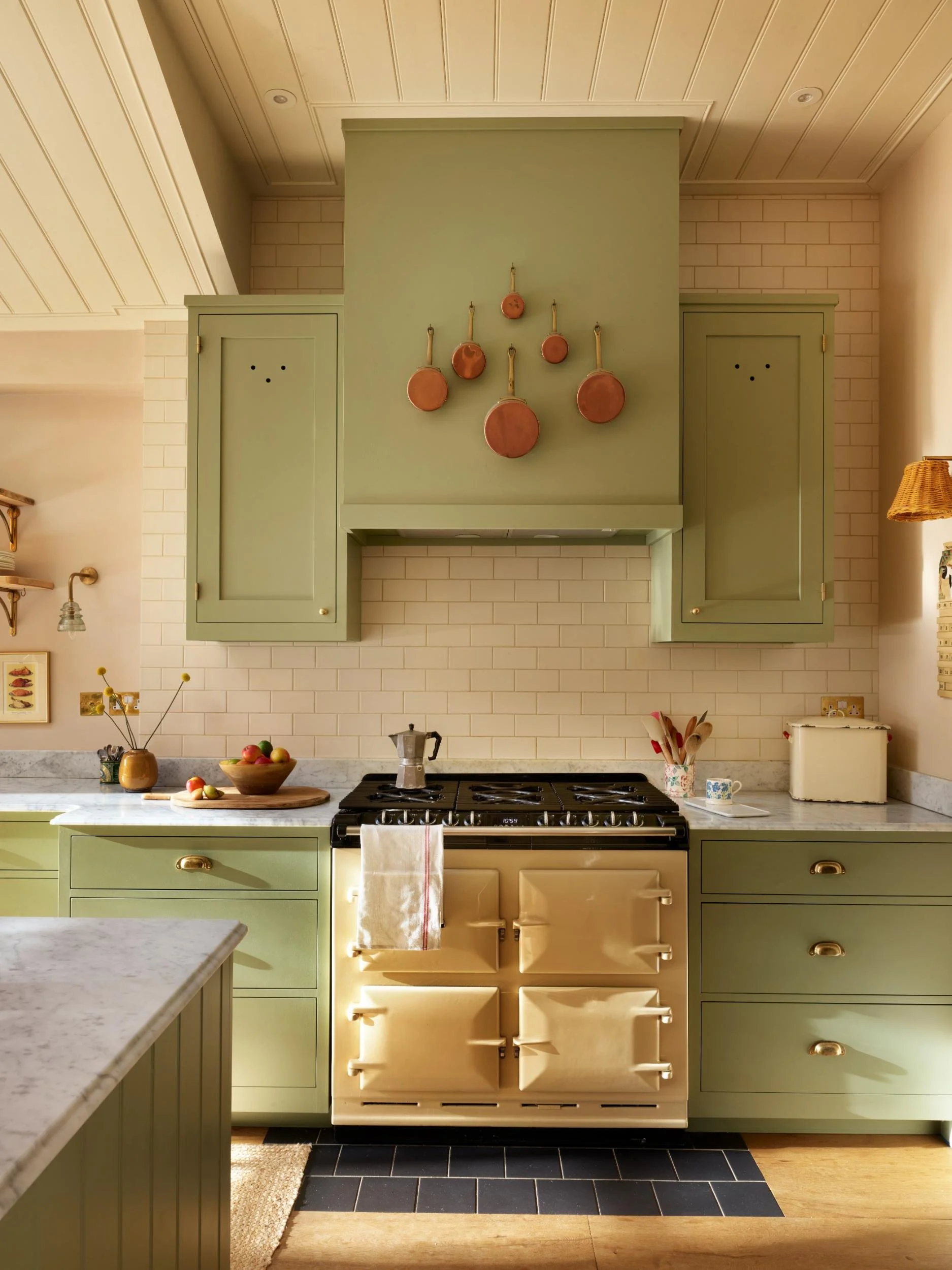 A cozy kitchen with green cabinets, a white oven, and a marble countertop. Copper pots are hanging on a green wall above the stove, with a bowl of apples on the counter.