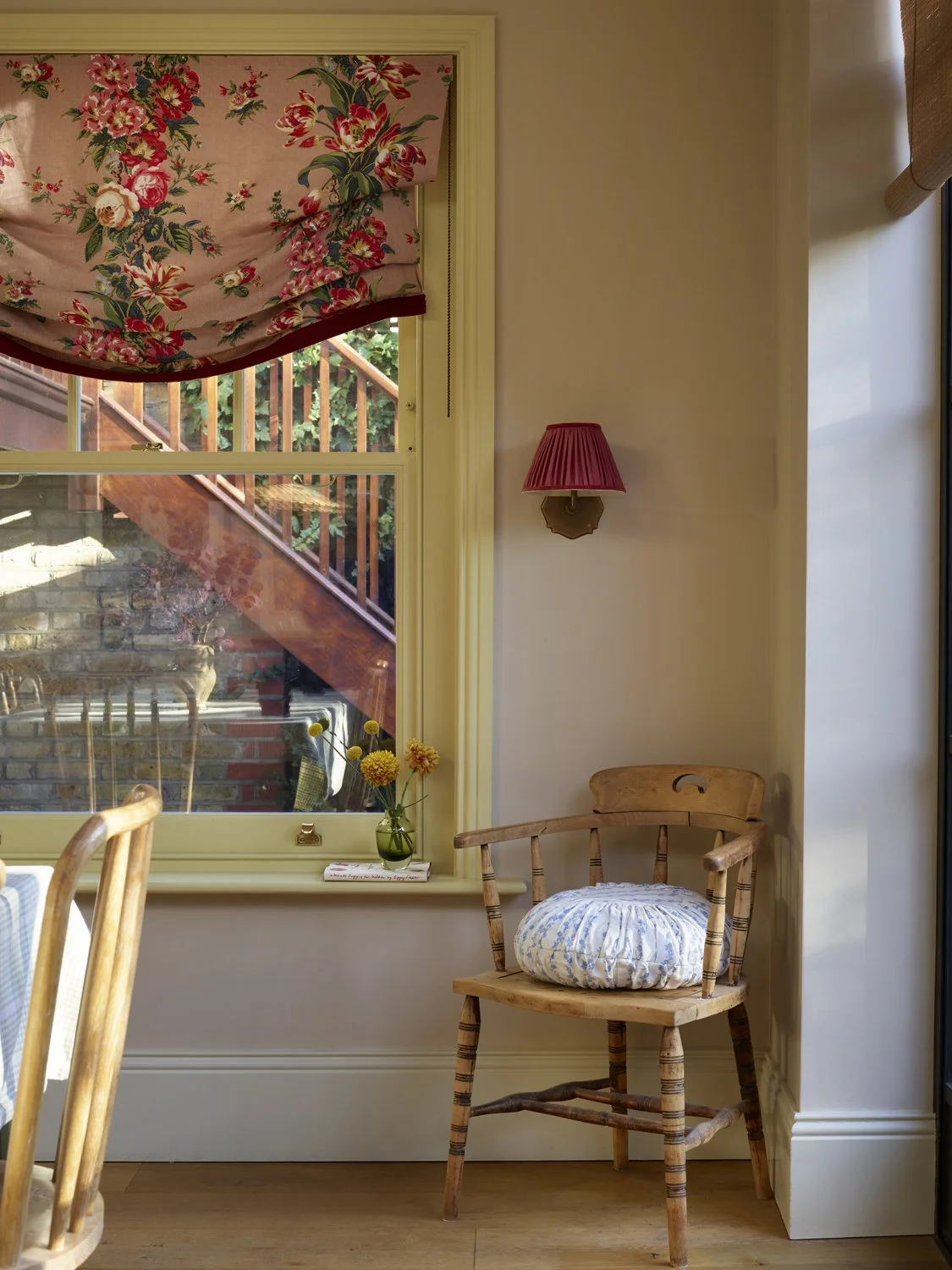A cozy corner with a wooden chair, a round pillow, a small red lampshade on the wall, a window with a floral curtain, a vase with yellow flowers, and stairs visible outside.