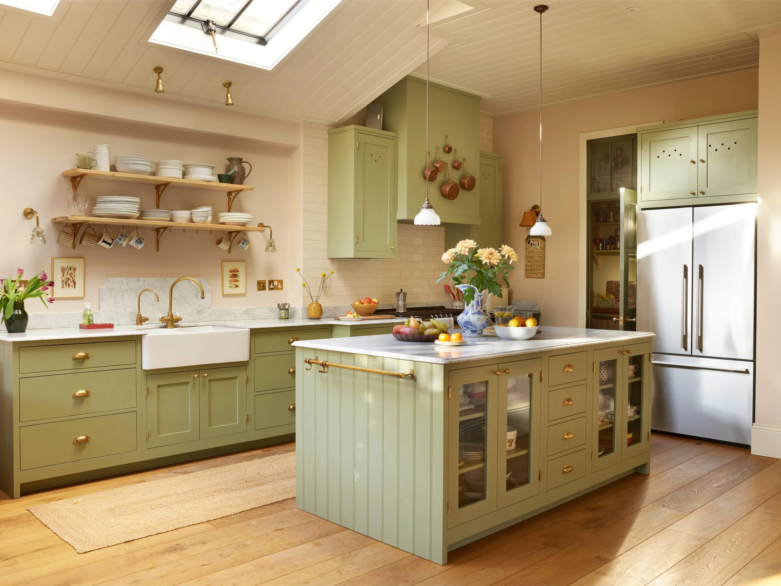 A kitchen with green cabinets and a large center island with a marble countertop, open shelving, hanging copper pots, a skylight, and various kitchen items and flowers on the island.