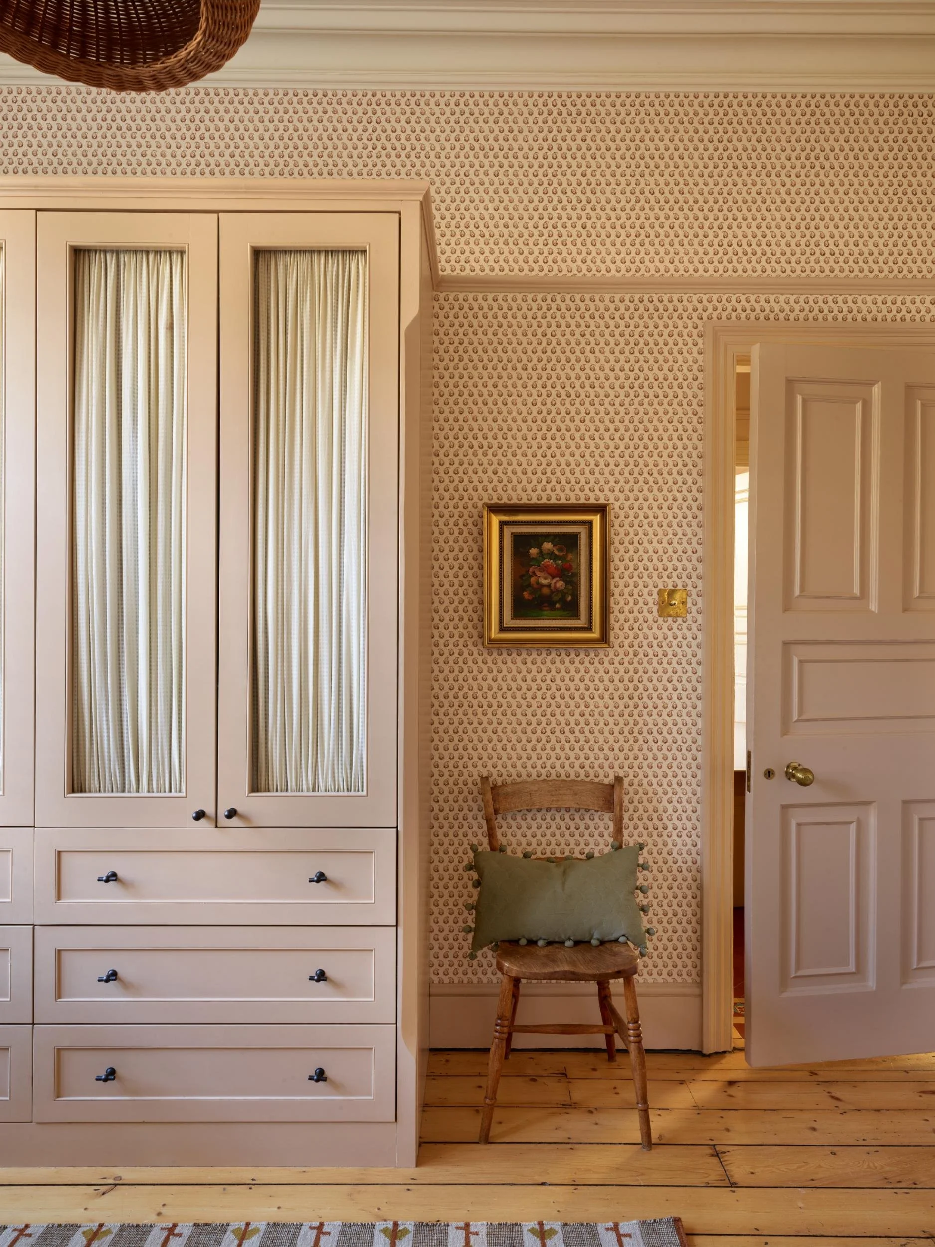A corner of a room with patterned wallpaper, a white wardrobe with glass-paneled doors, a small framed floral painting, a wooden chair with a green pillow, an open white door, and a woven basket hanging from the ceiling.