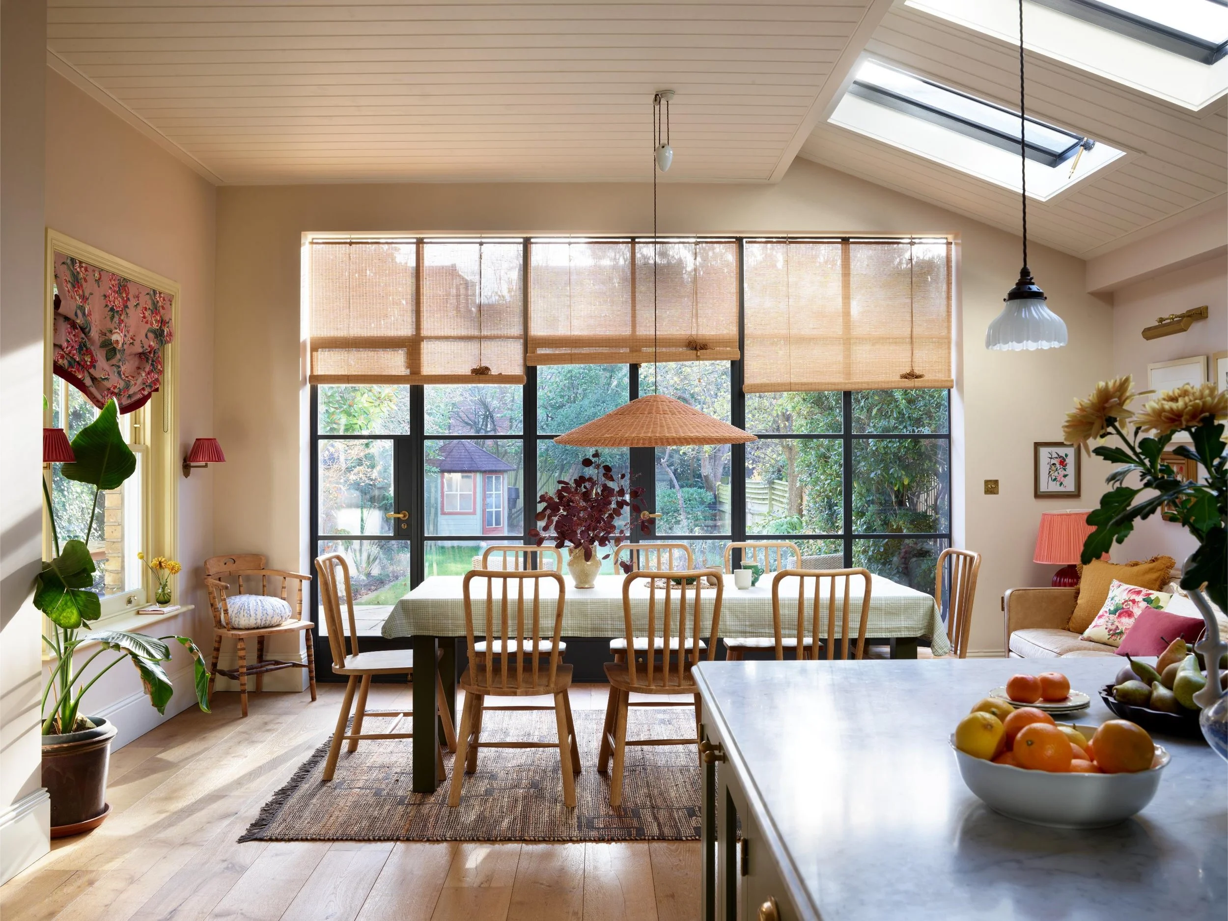 Bright kitchen or dining room with large windows, wooden kitchen table with chairs, plants, pendant lights, and a view of the backyard.