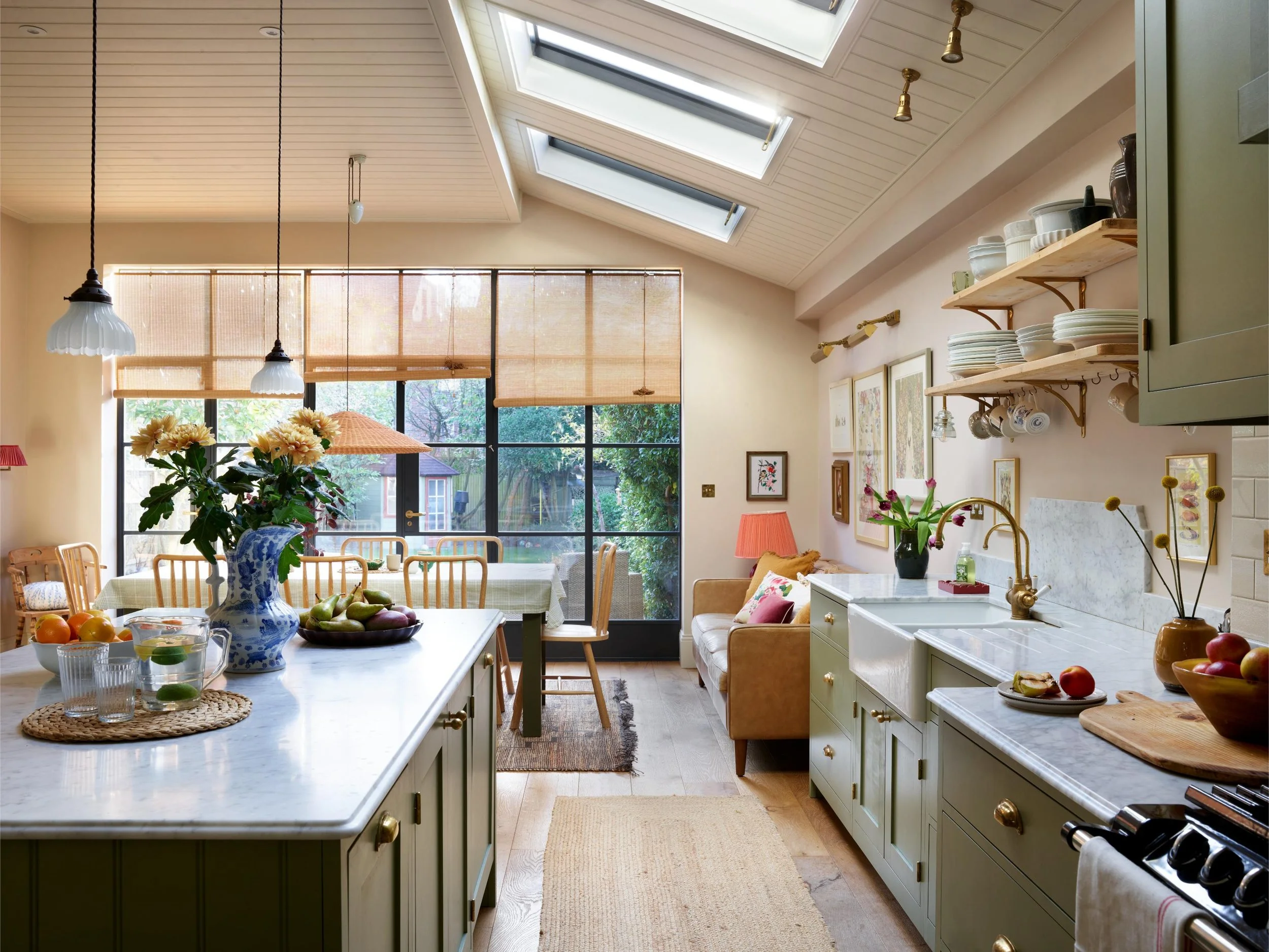 A bright kitchen with cream green cabinets, an island with food and drink, and a large window with a view of a backyard. Skylights and pendant lights hang from the ceiling.