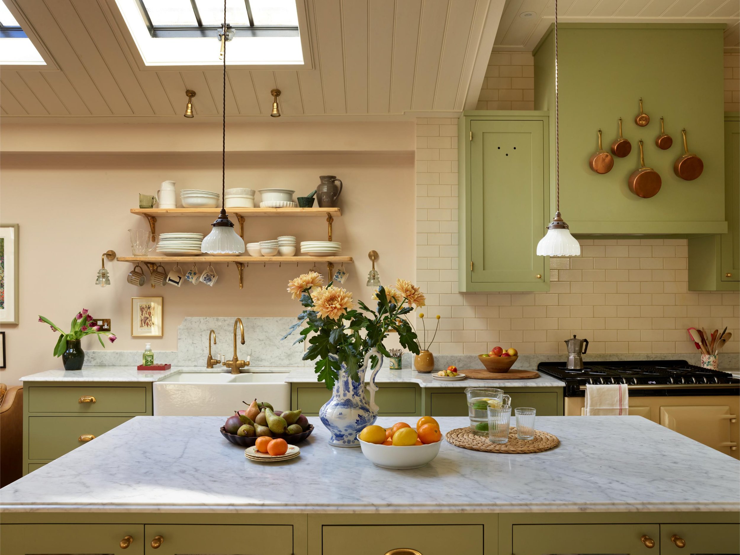 A bright kitchen with a white marble countertop, green cabinets, and open wall shelves holding dishes and bowls. There are copper pots hanging on the wall. A large window or skylight is above, and a hanging light fixture is shown. The kitchen island 