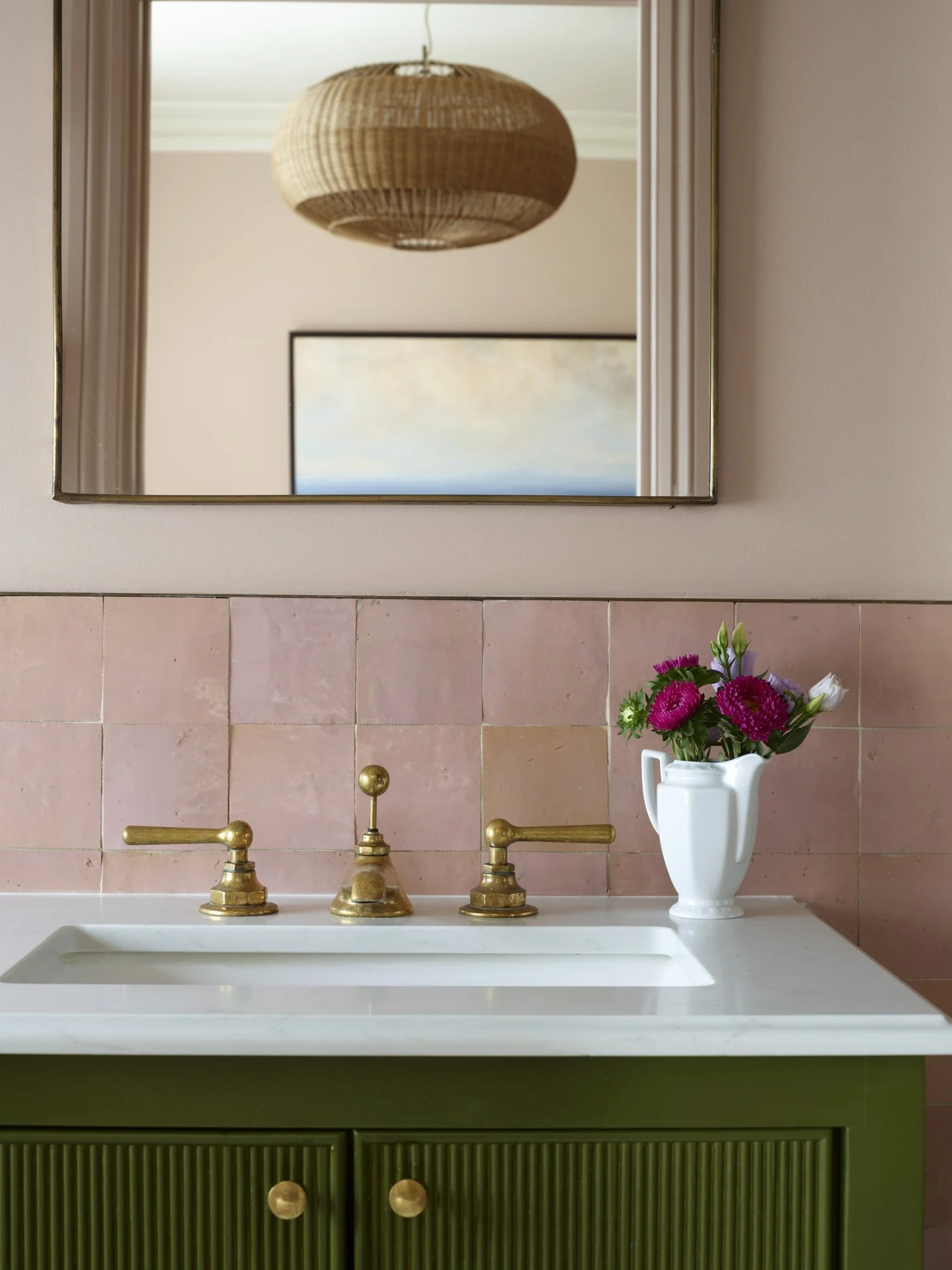 Bathroom with pink tiled wall, white sink, and brass faucet handles. Green cabinet with wooden knobs below the sink. White pitcher vase with pink, purple, and white flowers. A mirror above the sink reflects a wicker light fixture and a framed picture of a cloudy sky.