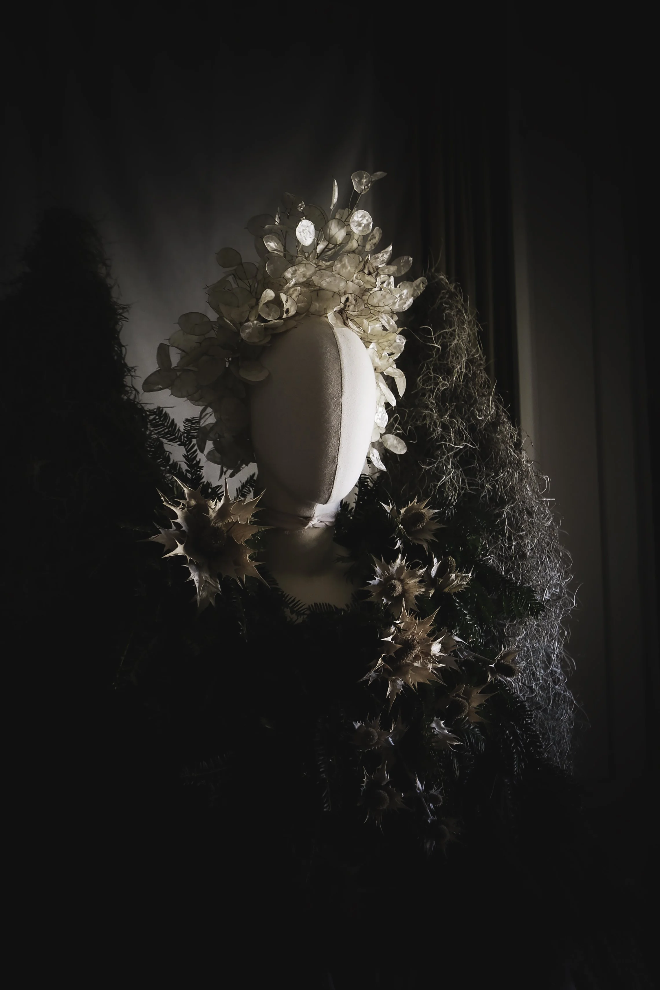 Decorative art piece with white leaves and flowers on a white mannequin head, surrounded by botanicals and greenery, with soft lighting creating shadows.