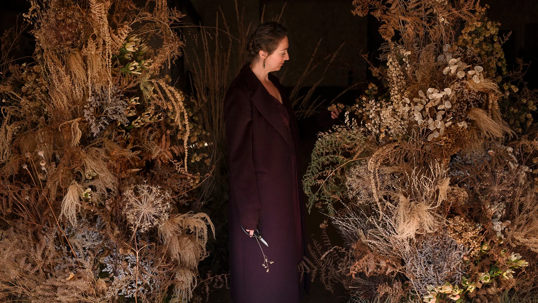 A woman in dark clothing standing among a backdrop of dried plants and flowers, holding a pair of scissors.