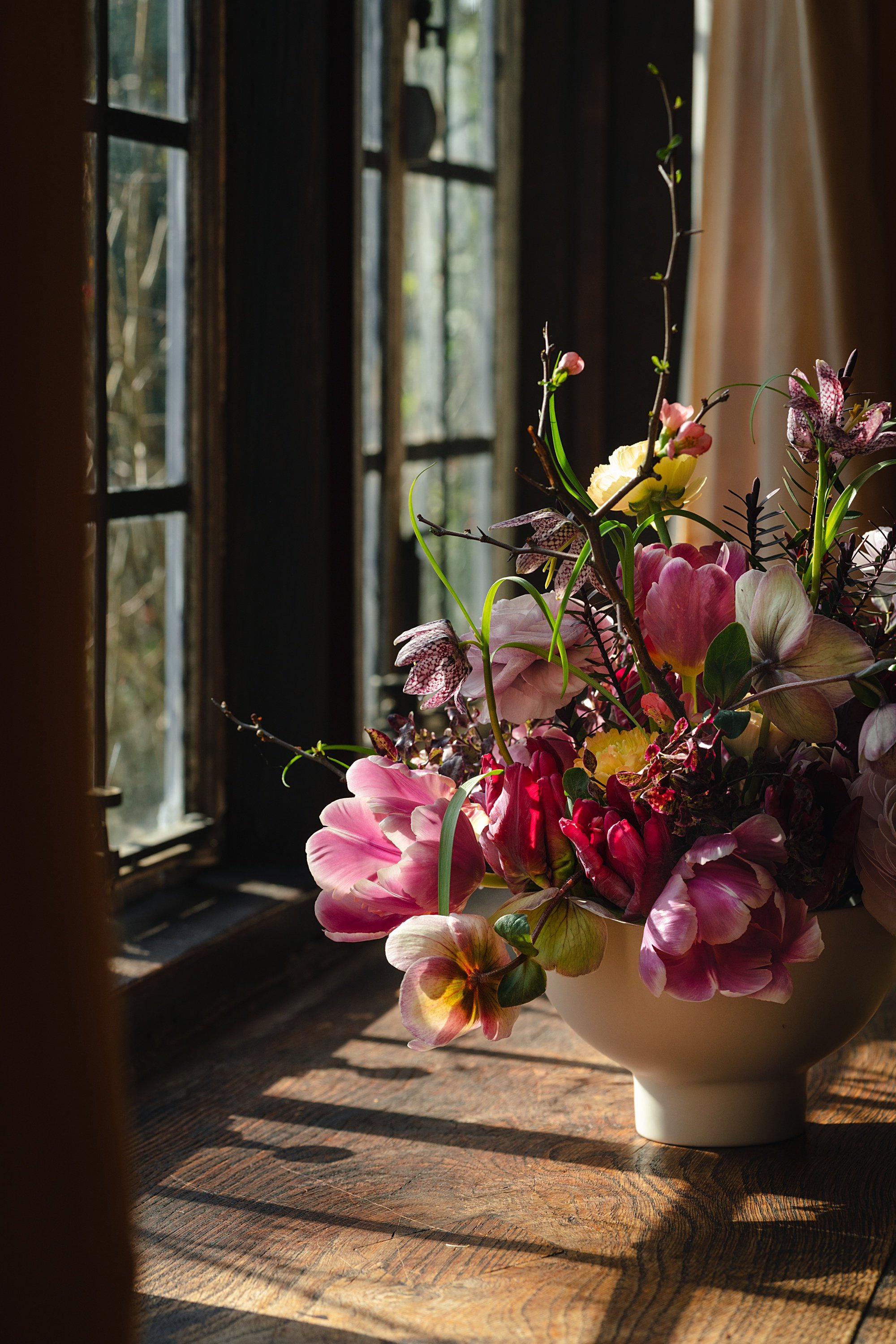 A colorful flower arrangement in a white vessel on a wooden table, near a window with sunlight streaming through.