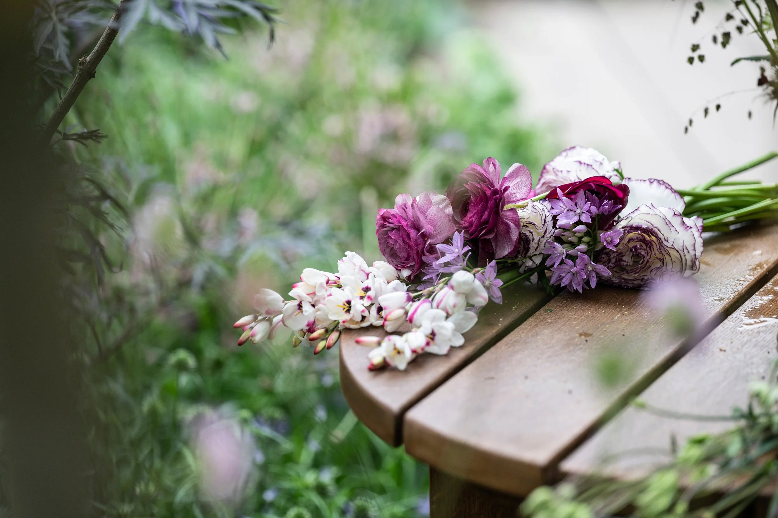 A wooden bench outdoors with a bouquet of pink, purple, and white flowers resting on it, surrounded by green foliage.