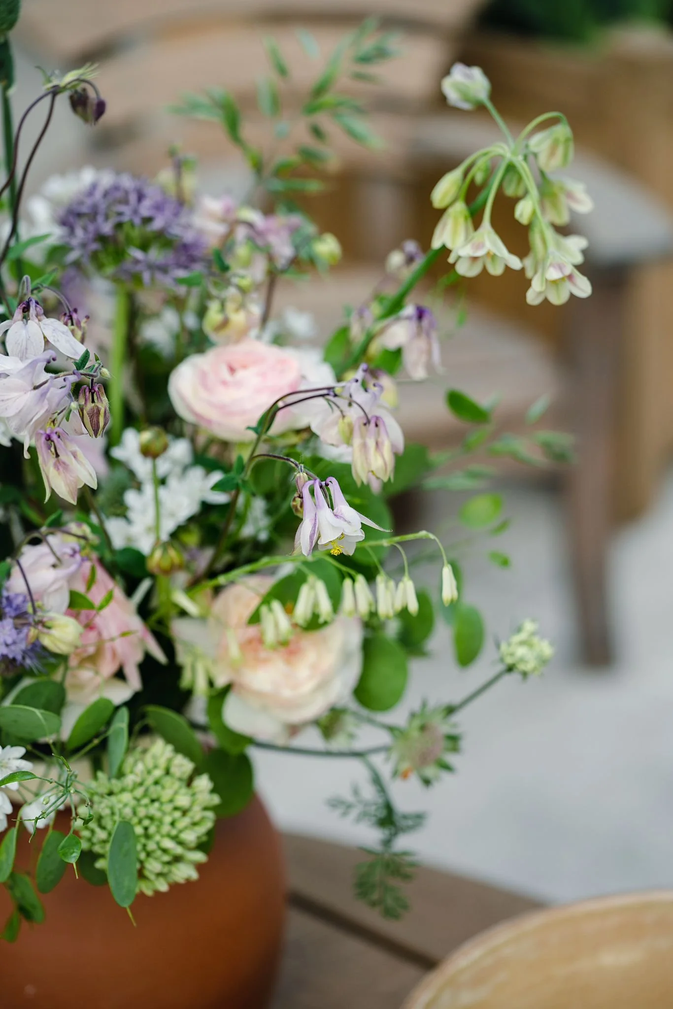 A close-up of a flower arrangement in a clay pot featuring purple, white, and pink flowers with green foliage on a wooden surface.