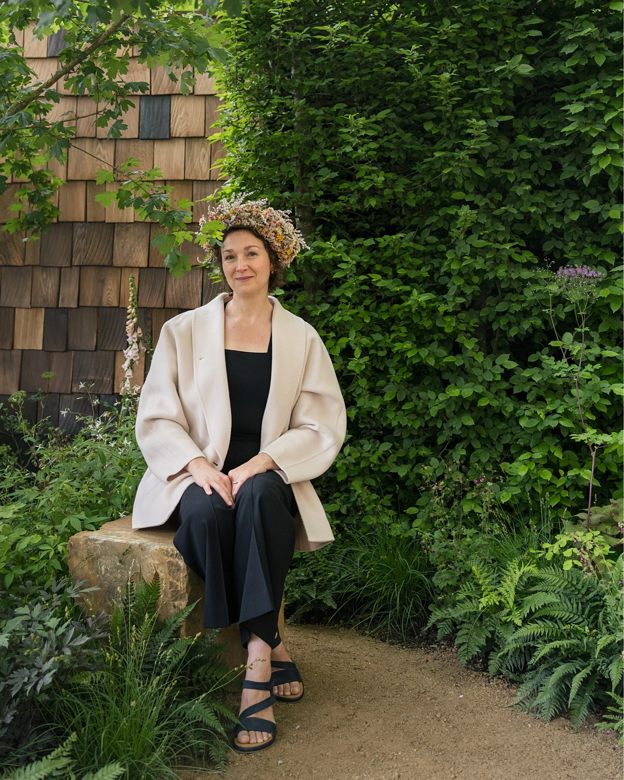 Woman sitting on a stone in a garden with green foliage and a wooden building in the background, wearing a floral crown, white blazer, black top, and black sandals.