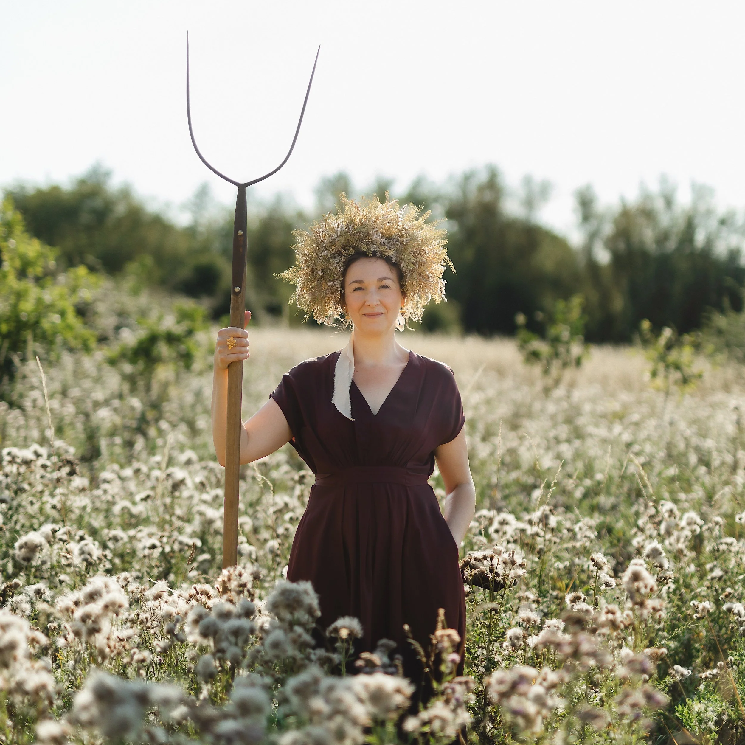 A woman wearing a floral crown standing in a field of blooming flowers, holding a pitchfork in her right hand, smiling at the camera.