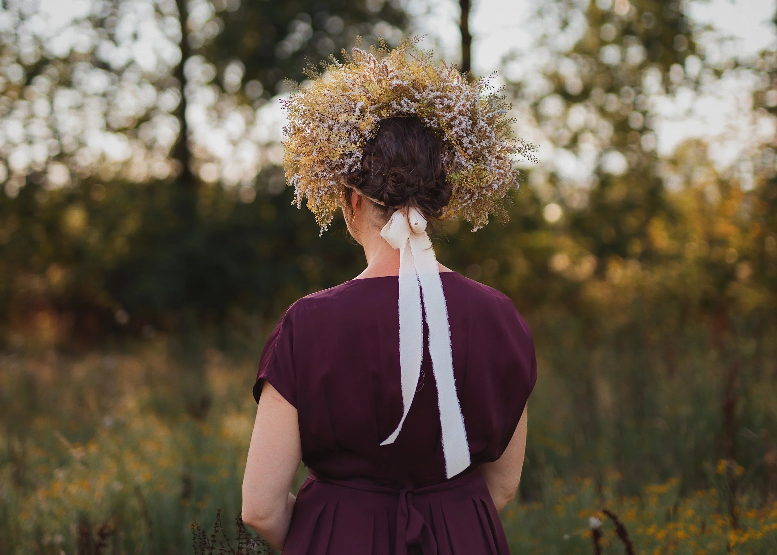 A woman from behind with curly hair wearing a floral crown made of yellow, white, and pink flowers, standing outdoors in a wooded area during autumn.