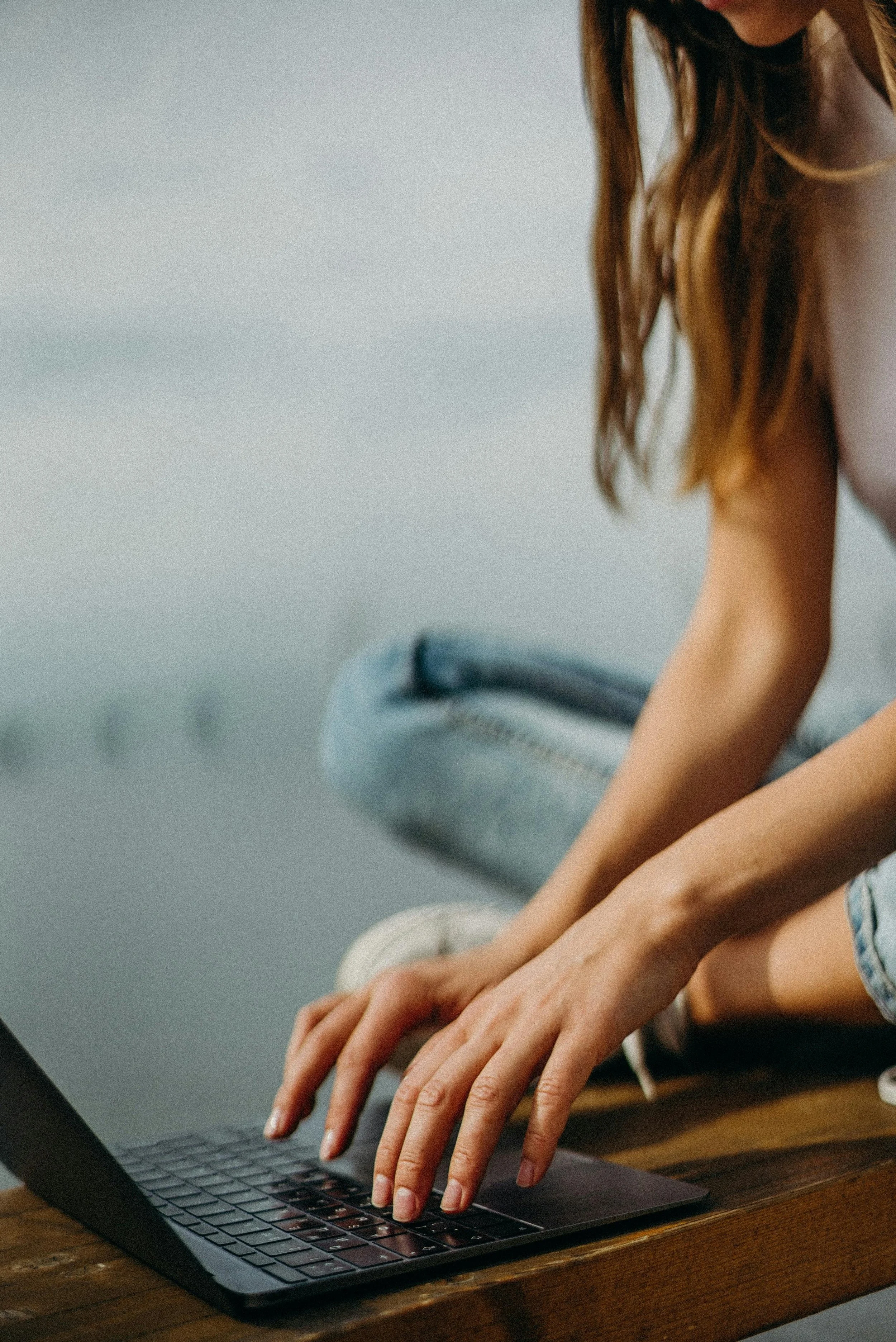 Close-up of a woman typing on a laptop keyboard, sitting outdoors on a wooden surface, wearing a white shirt and jeans.