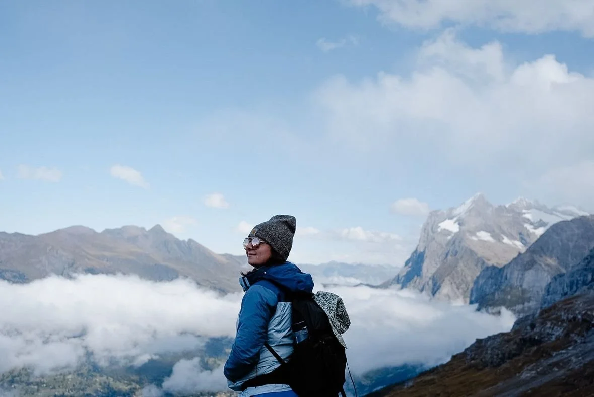 A person wearing a gray beanie, sunglasses, and a blue jacket with a backpack, standing outdoors in a mountainous landscape with clouds and snow-capped peaks.