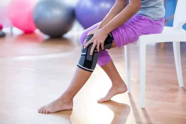 A person sitting on a white chair using a knee brace in a gym or rehabilitation center with gym balls in the background.