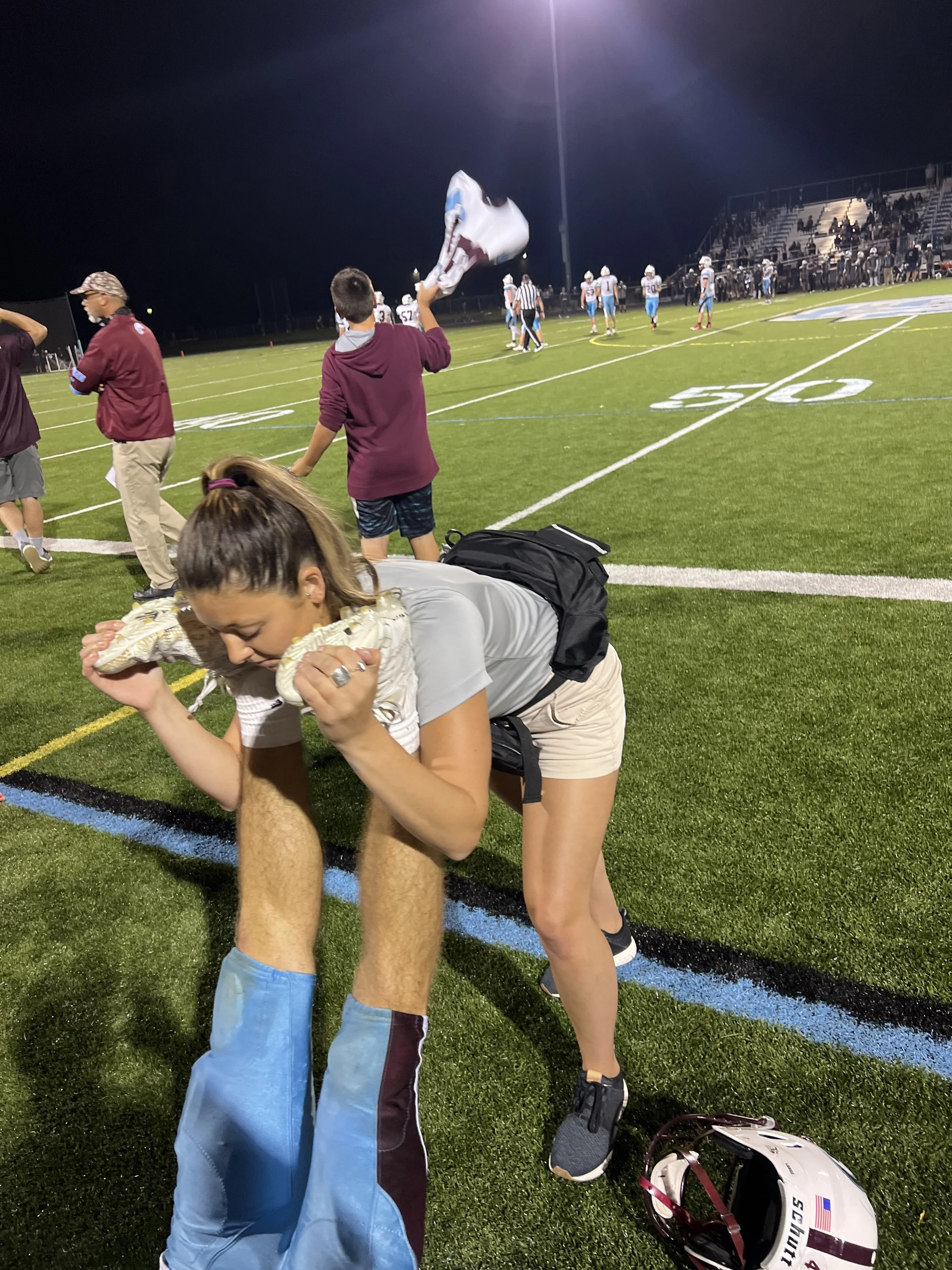 A woman dressed in athletic clothing holds a football helmet. She is kneeling and leaning on a person's legs, who is lying on the ground with their legs upward. The scene takes place on a football field at night, with players and spectators in the background.