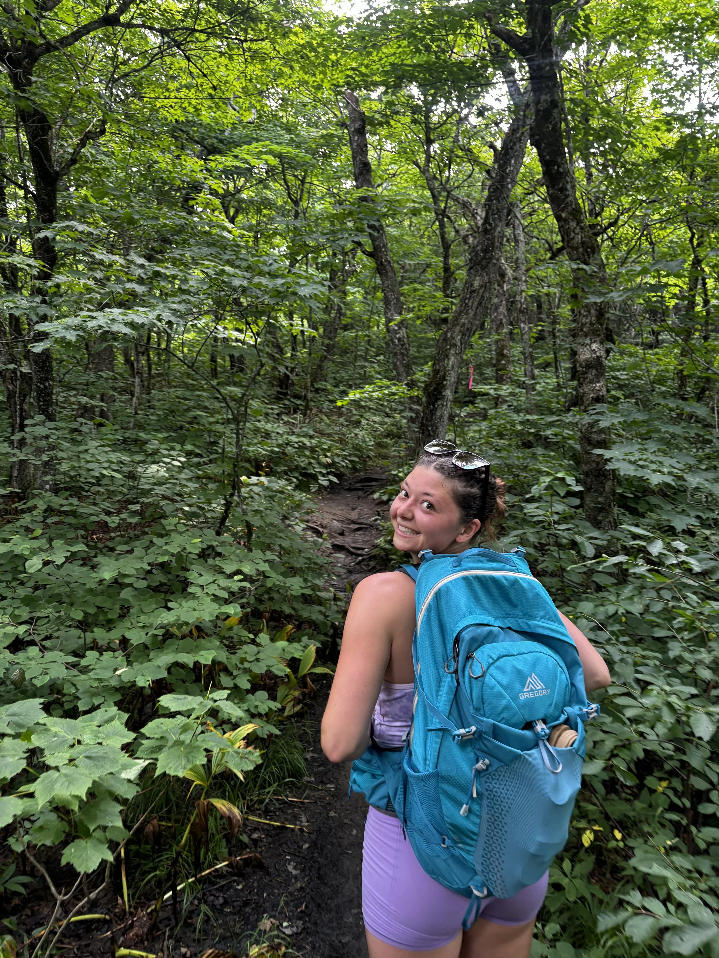 A young woman with a blue backpack smiling while hiking on a narrow dirt trail through a green forest.