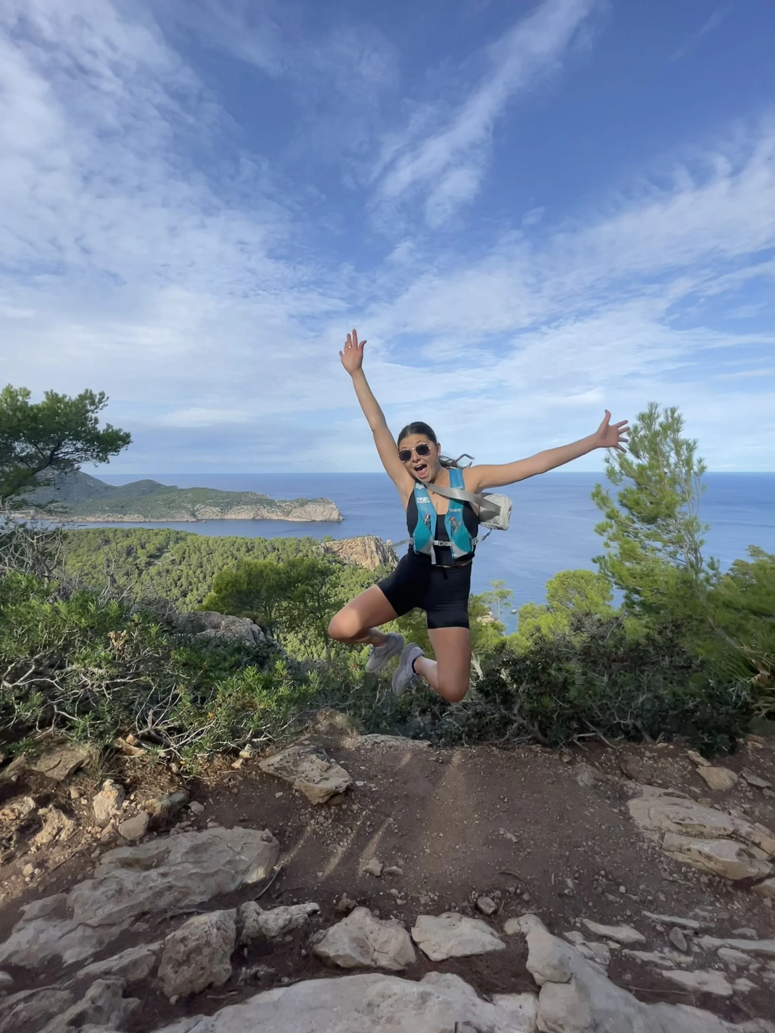 An excited woman jumping on a hiking trail with a scenic view of the ocean, cliffs, and blue sky in the background.