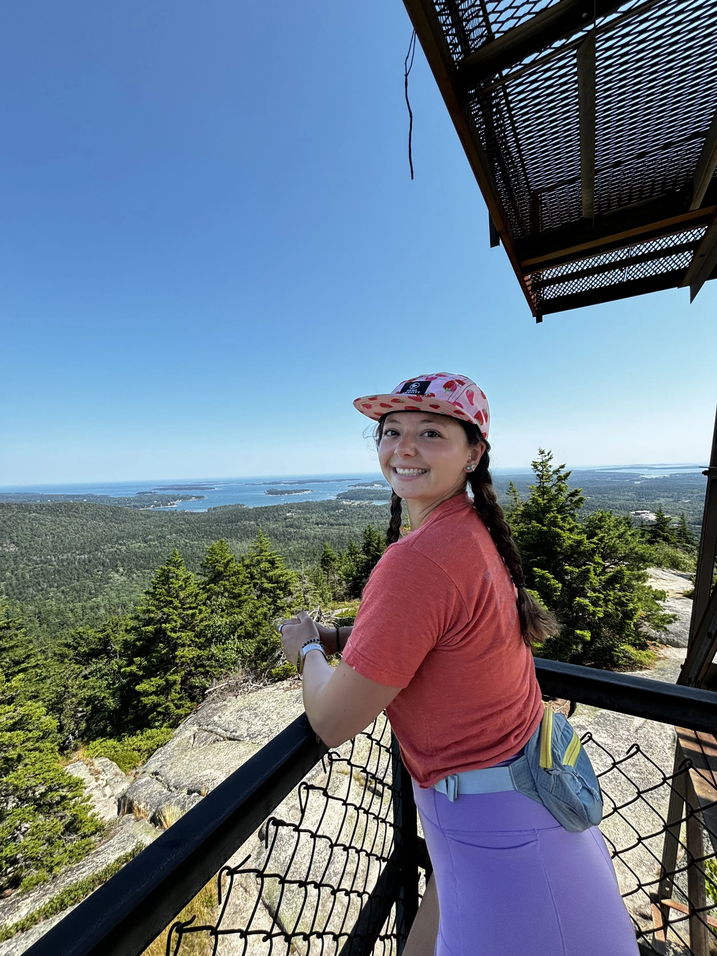 A young woman with braided hair smiling on a wooden observation deck overlooking a vast forest and distant lakes under a bright blue sky.