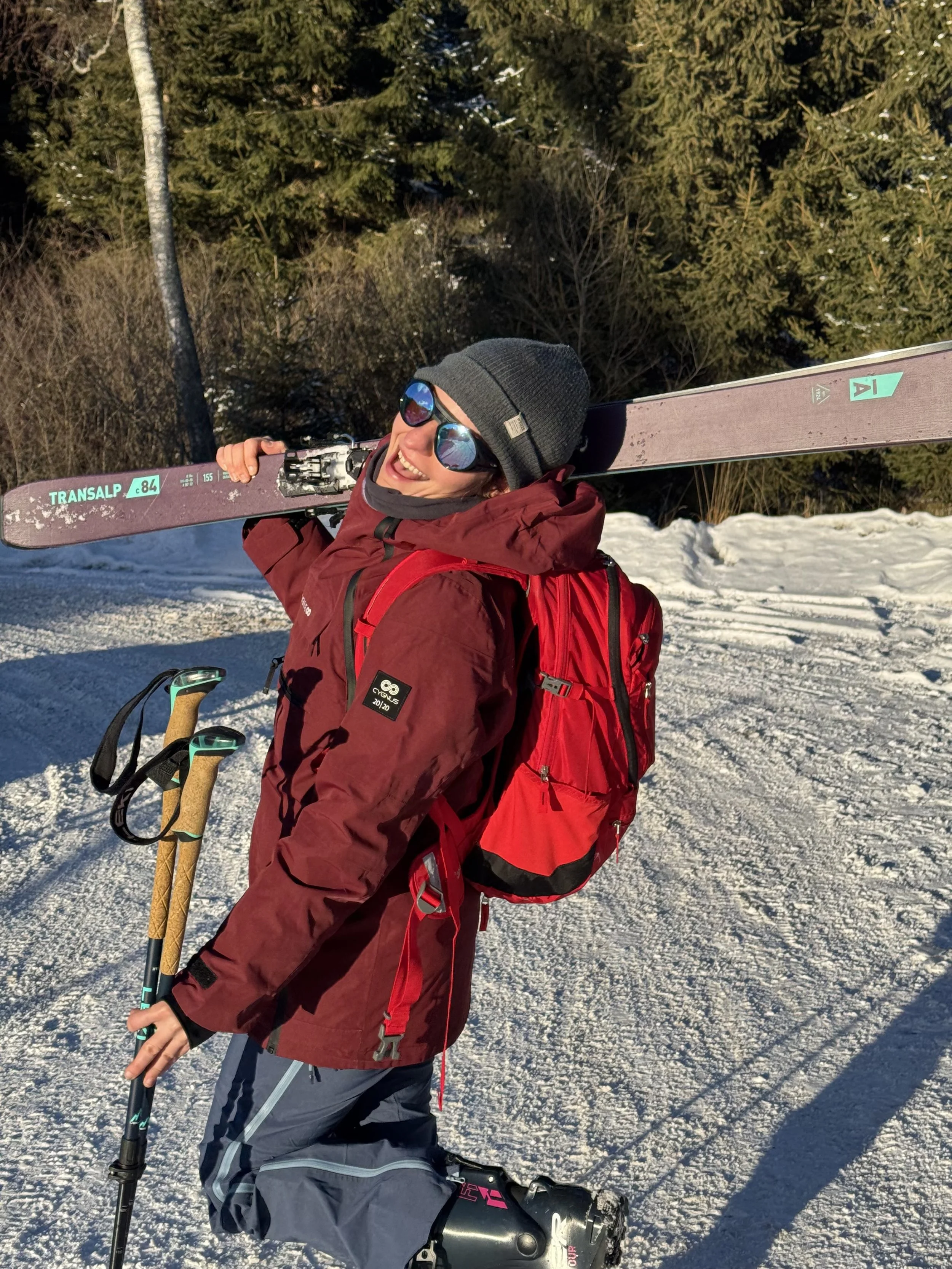 Lächelnde Skifahrerin mit roter Jacke, Rucksack, Skiern und Sonnenbrille im verschneiten Wald