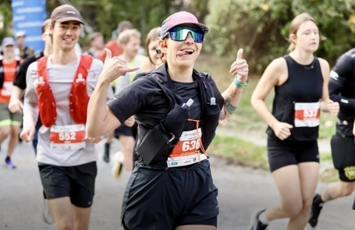 Läuferinnen und Läufer bei einem Straßenlauf im Park; eine Läuferin mit Sonnenbrille und Startnummer 636 zeigt eine lockere Geste.