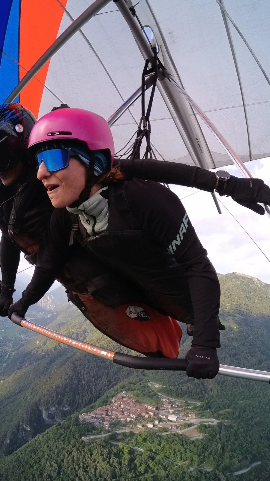 Zwei Personen beim Drachenfliegen über einer bewaldeten Berglandschaft mit Dorf im Tal